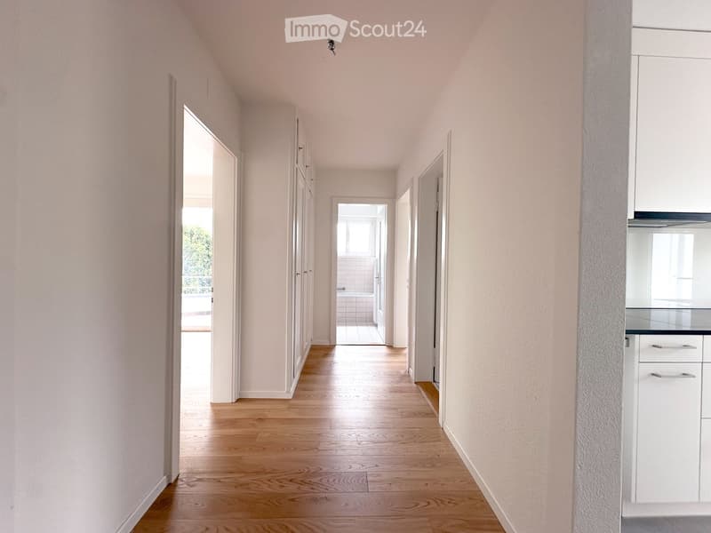 A long, narrow hallway with light wooden flooring and white walls, leading to several rooms including a kitchen and a bathroom.