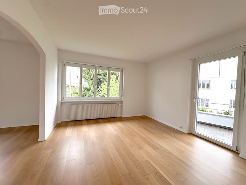 Empty room with wooden flooring, large windows, and a sliding glass door leading to a balcony.
