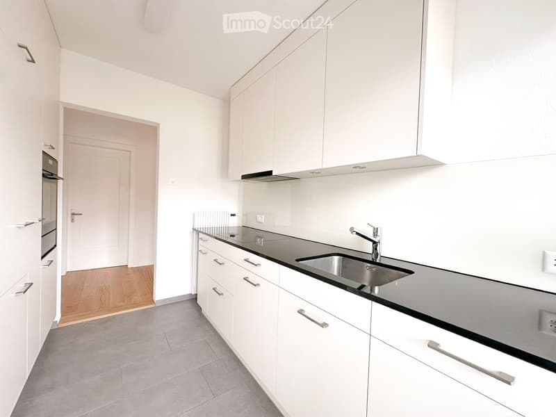 Modern kitchen with white cabinets, stainless steel sink, and a built-in oven. The space features a sleek, minimalist design with a black countertop and grey tiled floor.