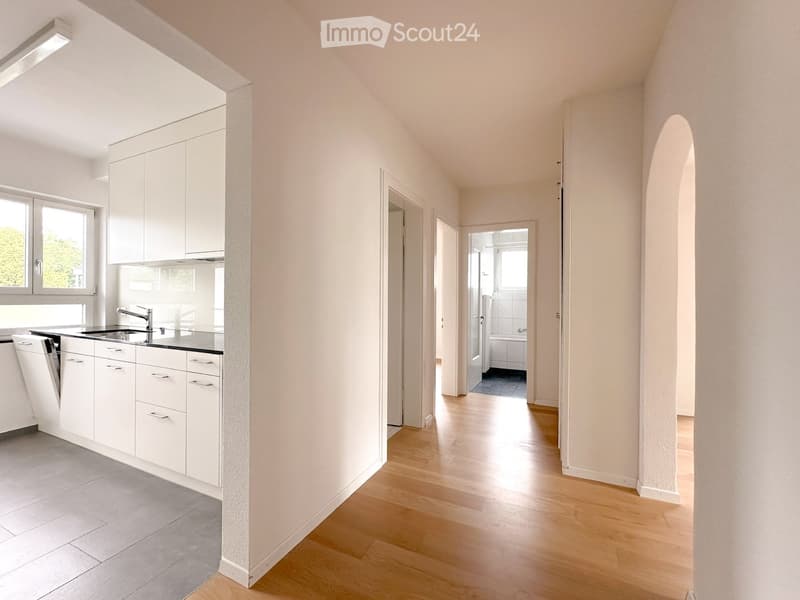 Modern kitchen with white cabinets, a sink, and a dishwasher, connected to a hallway leading to a bathroom.