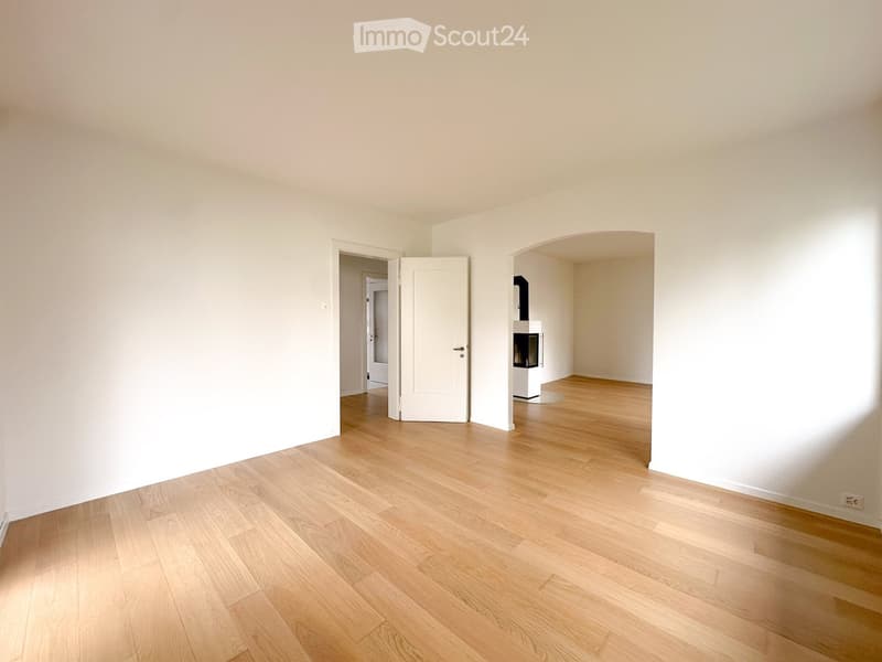 Empty room with light wooden flooring and white walls, featuring an open doorway leading to another room and a fireplace in the adjacent space.