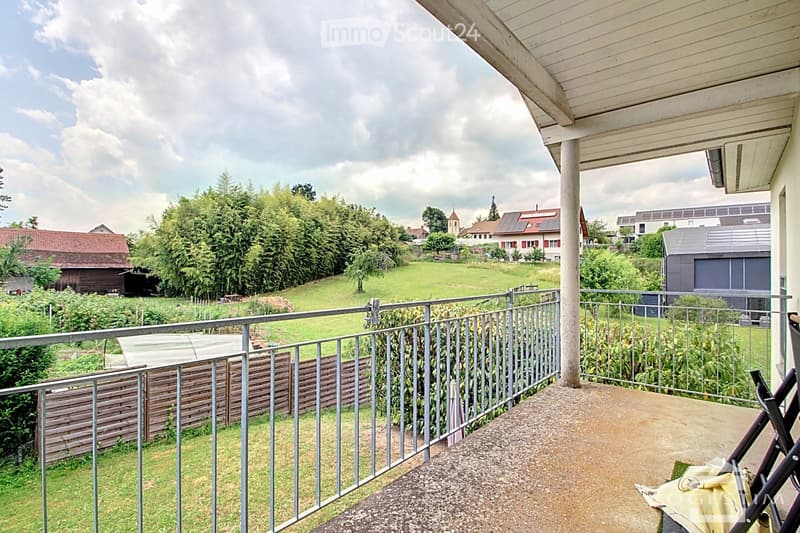 Terrace with metal railing, a house behind, and green area with plants and grass