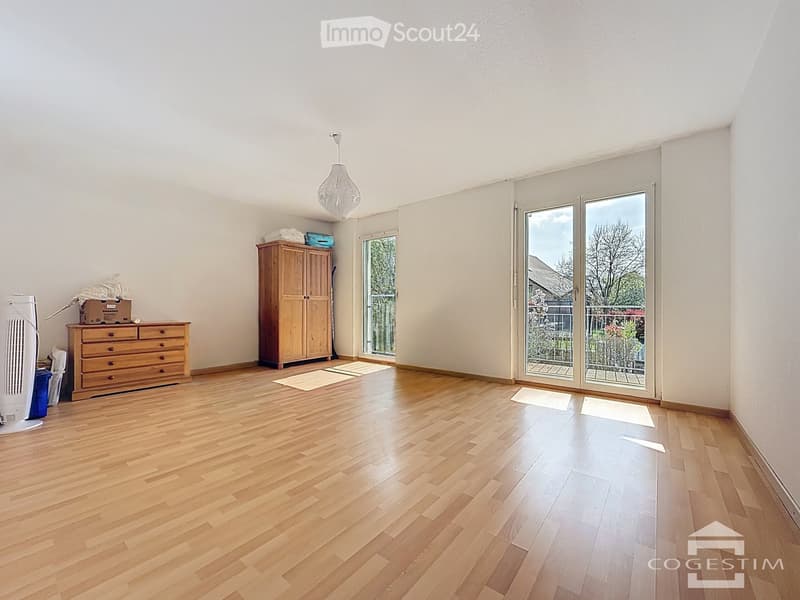 Bright room with wooden flooring, large windows, and sliding glass doors leading to a balcony. Contains a wardrobe, chest of drawers, fan, and a ball lamp.