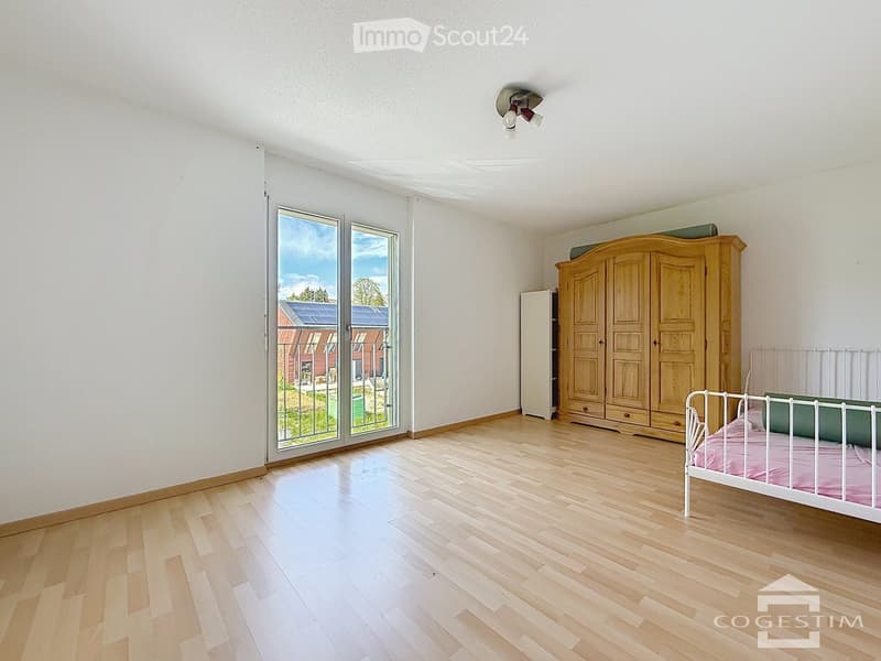 Bright bedroom with wooden flooring, large windows, a single bed with pink bedding, a wooden wardrobe, and a small cabinet.