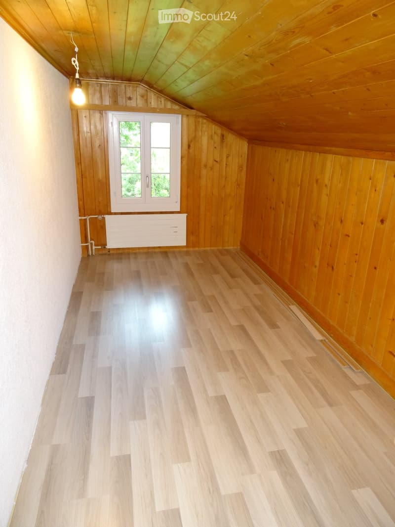 Empty attic room with wooden walls, wooden floor, and a single window with white frame. The room has a slanted wooden ceiling and a built-in radiator below the window.
