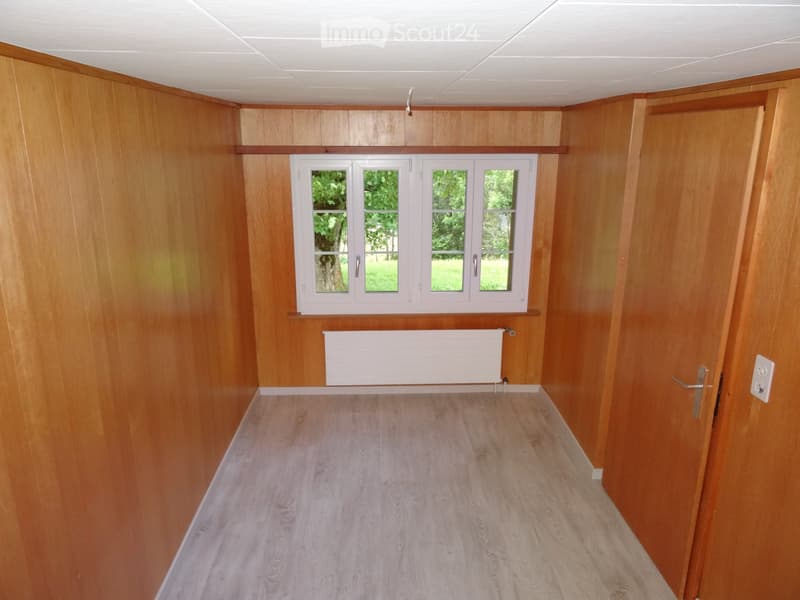 Empty room with wooden walls, light wooden flooring, and a white radiator beneath a window with four panes. The room has a door on the right side.