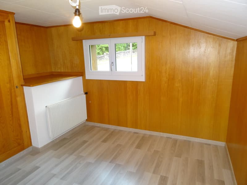 Empty room with wooden walls, wooden floor, white radiator, window with white frame, and a built-in shelf.