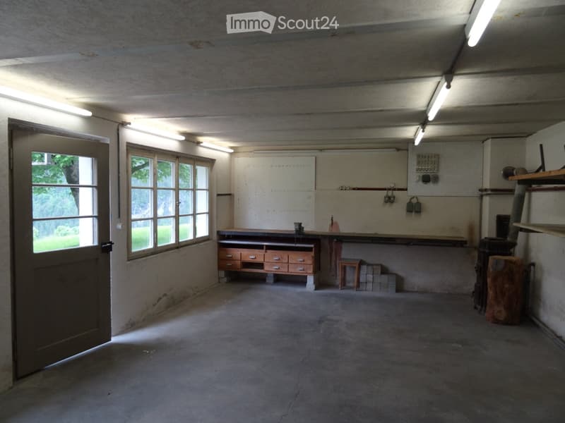 Workshop or garage space with concrete floors, large windows, a workbench, and various tools and storage items.