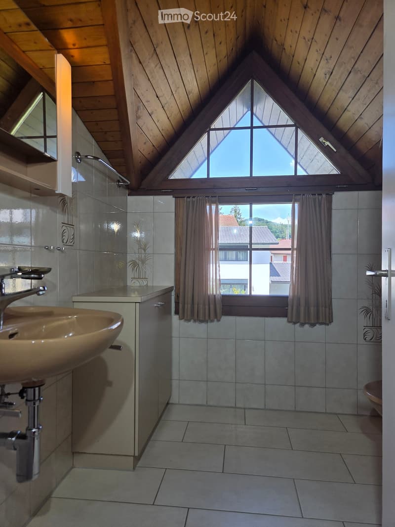 Bathroom with a sink, faucet, cabinet, and a shower area. The room features wooden ceilings and large windows with curtains, allowing natural light to fill the space.