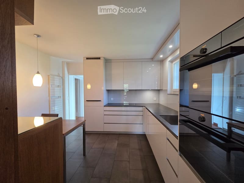 Modern kitchen with white cabinets, dark countertops, and built-in appliances including an oven. Features a wooden dining table and pendant lighting.