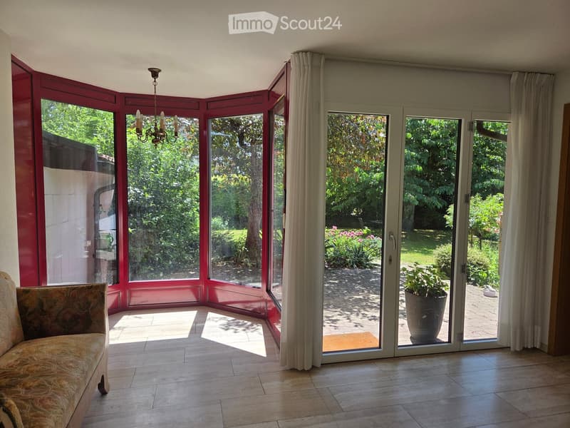Bright living room with large windows and sliding glass doors opening to a garden, featuring a patterned sofa and a chandelier.