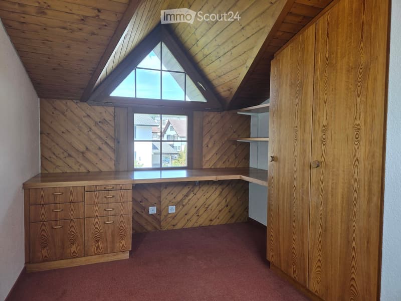 An empty attic room with wooden walls, a slanted wooden ceiling, and a large skylight window. Features a built-in wooden desk with drawers and a small shelf unit.