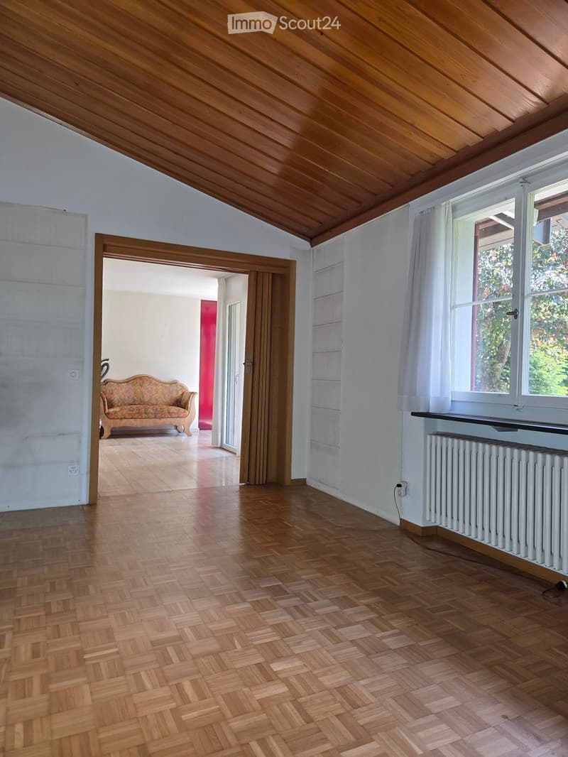 Bright, wooden-floored room with slanted wooden ceiling, large window with white curtains, radiator, and an open doorway leading to another room with a patterned sofa.