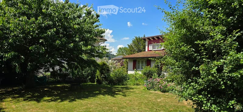 A well-maintained garden with a lawn, trees, and a small table and chairs set under the shade of a large tree. A red house with white trim is visible in the background.