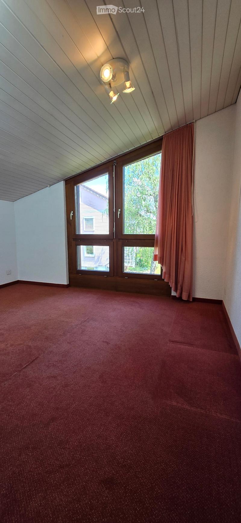 Empty room with red carpet, wooden door, large windows with curtains, and a slanted ceiling with built-in lighting.