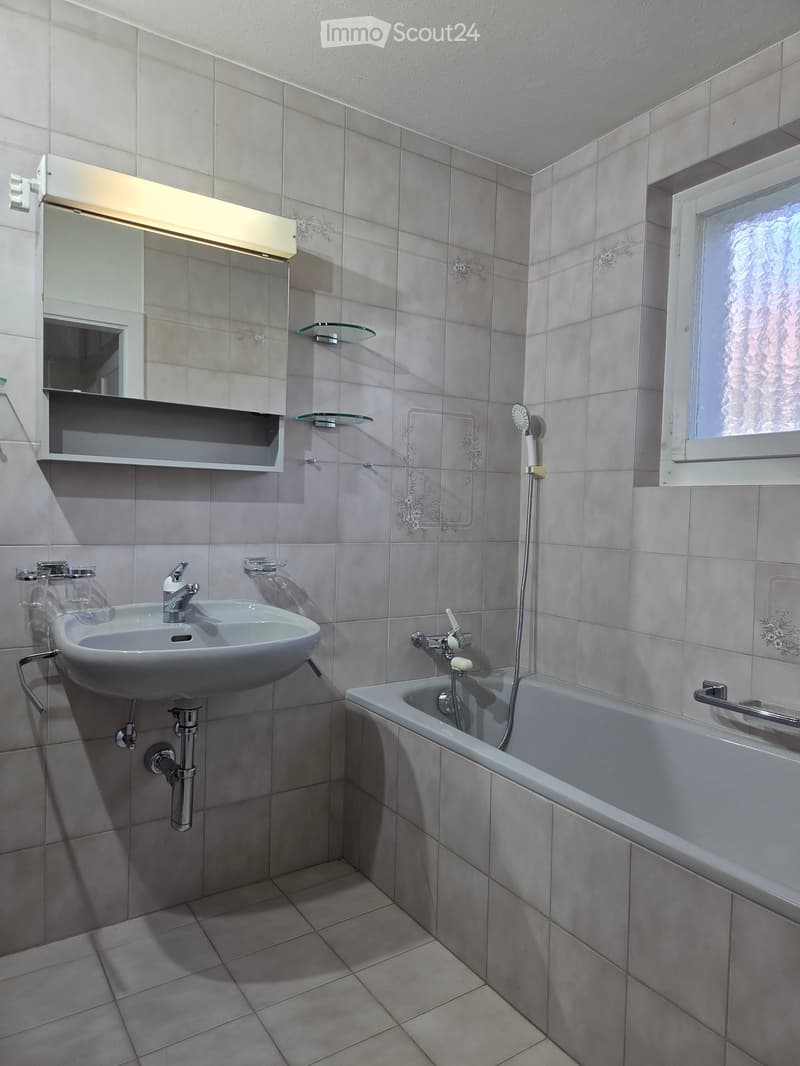 Modern bathroom with a white sink, bathtub, mirror with built-in storage, and glass shelves. The walls are tiled in a light grey color with decorative floral patterns.