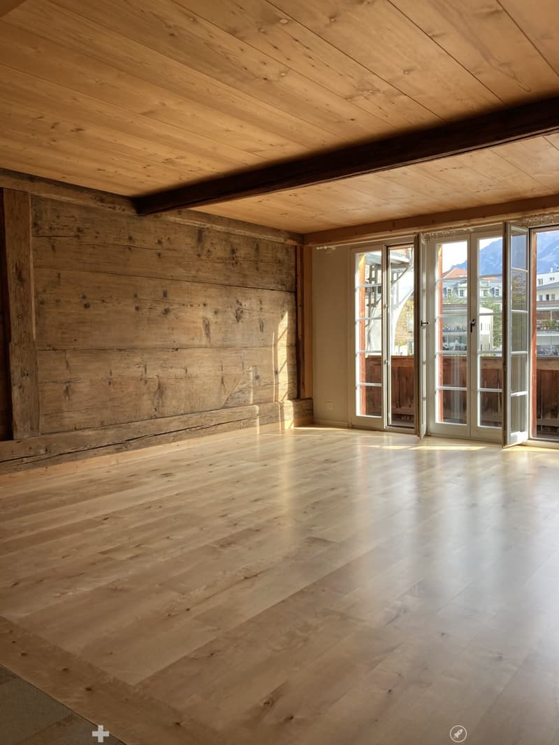 Empty room with wooden floors and exposed wooden beams on the ceiling. Large windows and doors provide natural light and access to the outdoors.