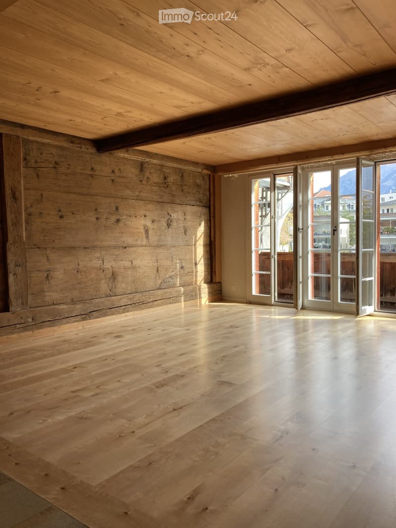 Empty room with wooden floors and exposed wooden beams on the ceiling. Large windows and doors provide natural light and access to the outdoors.