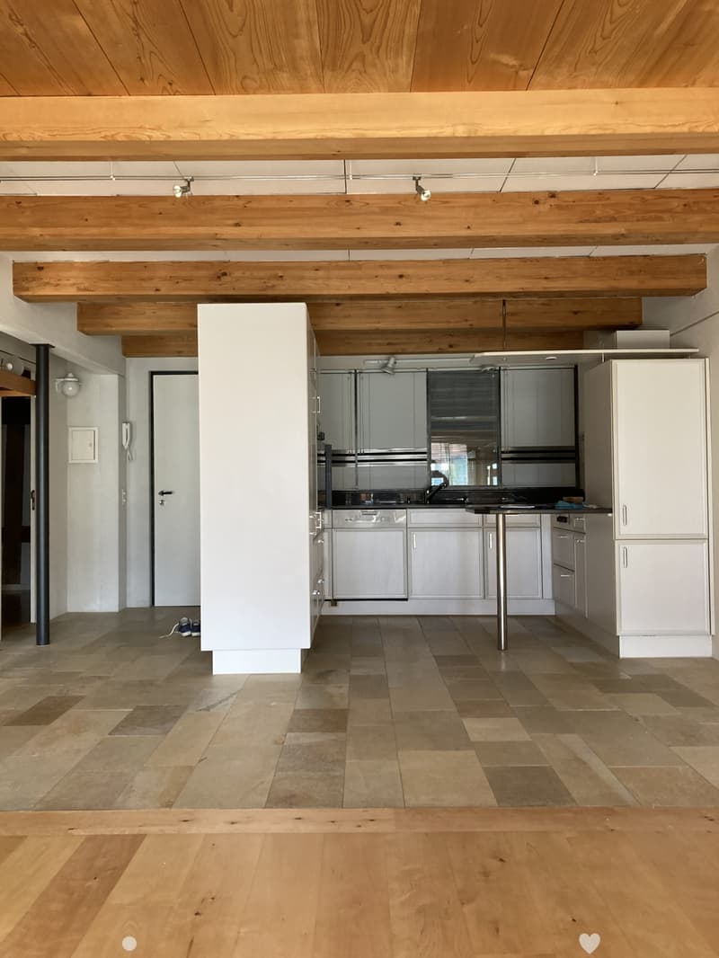 Modern kitchen with white cabinets, stainless steel appliances, and a central island. Exposed wooden beams on the ceiling and a mix of tiled and wooden flooring.