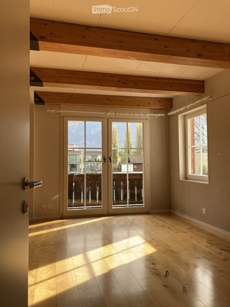 Empty room with wooden flooring, exposed wooden beams on the ceiling, large sliding glass doors leading to a balcony, and a window on the right side.