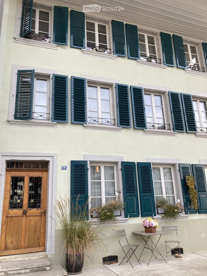 A two-story building with light yellow walls, green shutters, and multiple windows. The ground floor features a wooden door, a small table with chairs, and potted plants, while the upper floor has several windows with shutters and small flower boxes.