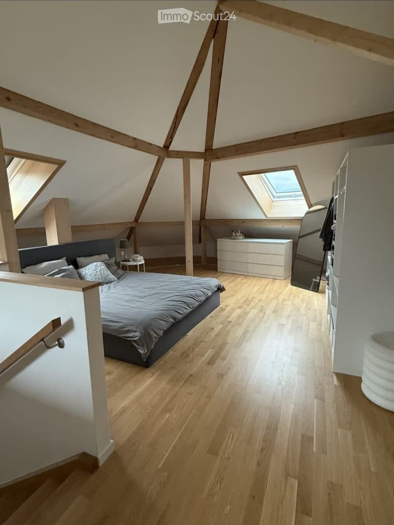 Bright, modern attic bedroom with wooden floors, slanted wooden ceiling beams, skylight windows, and contemporary furnishings including a bed, nightstand, and dresser.