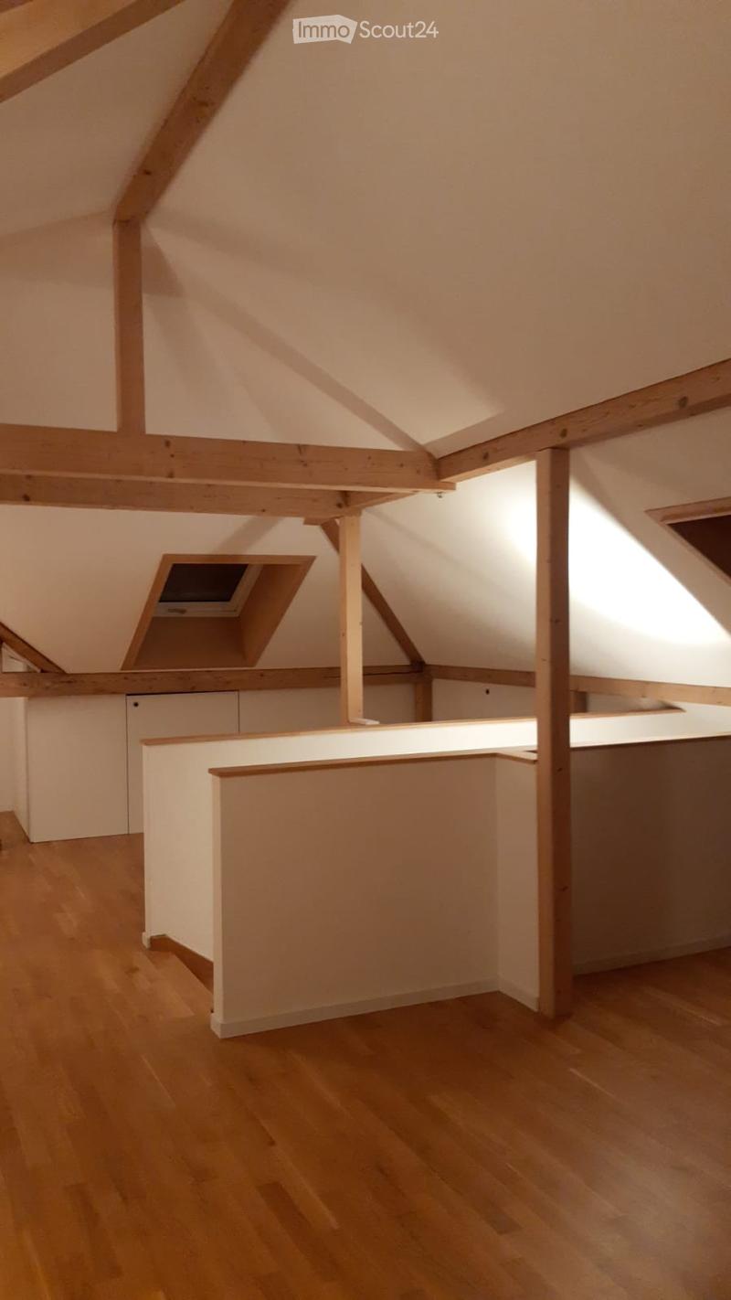An attic space with wooden beams, exposed wooden floor, and built-in cabinets. The room features a skylight window and a small bar counter area.