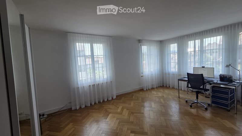 Bright, modern office space with herringbone wooden flooring, large windows with sheer curtains, a desk with a computer, a chair, and a desk lamp.
