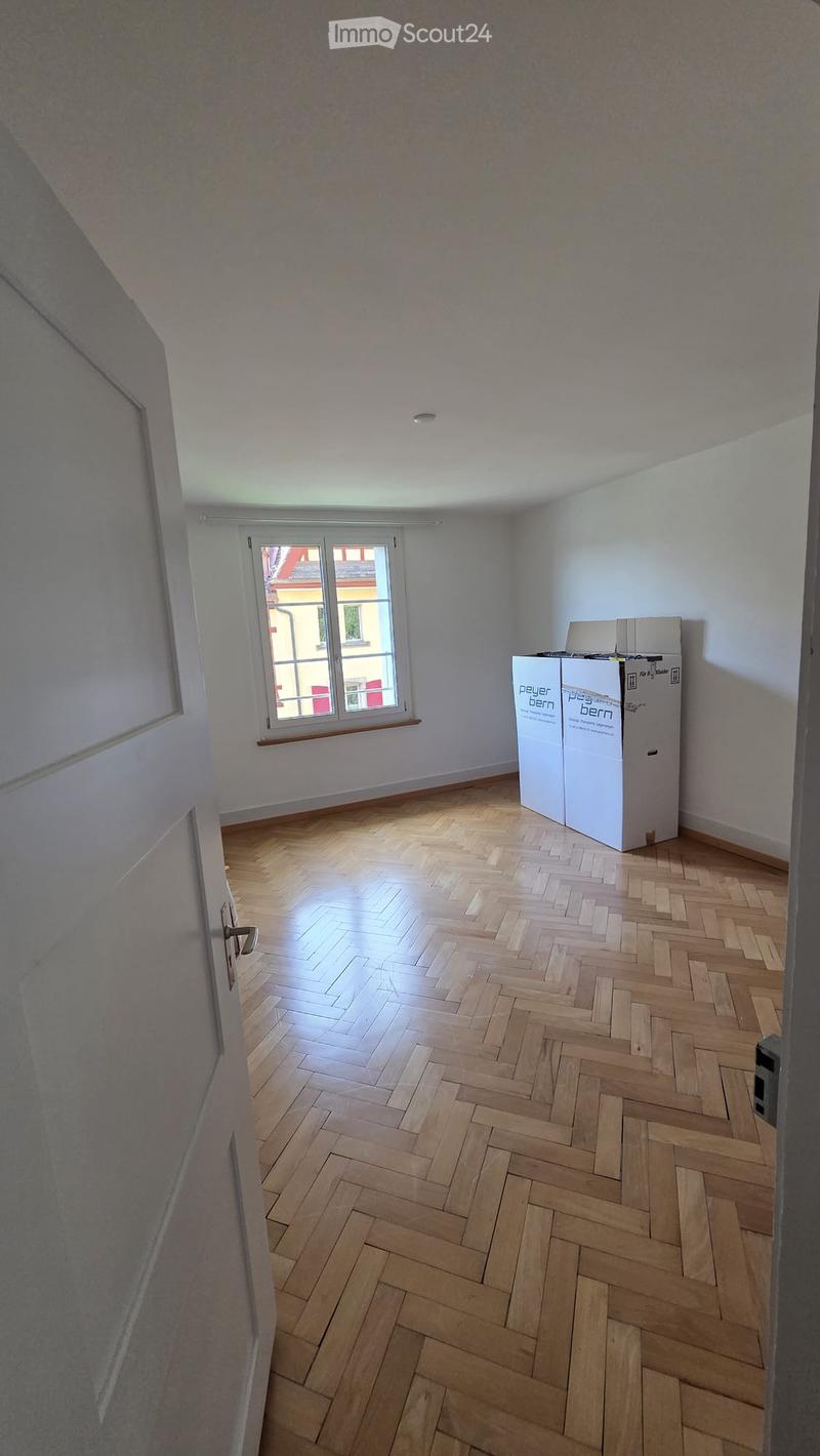 Empty room with wooden herringbone flooring, white walls, and a window with a view of a neighboring building.