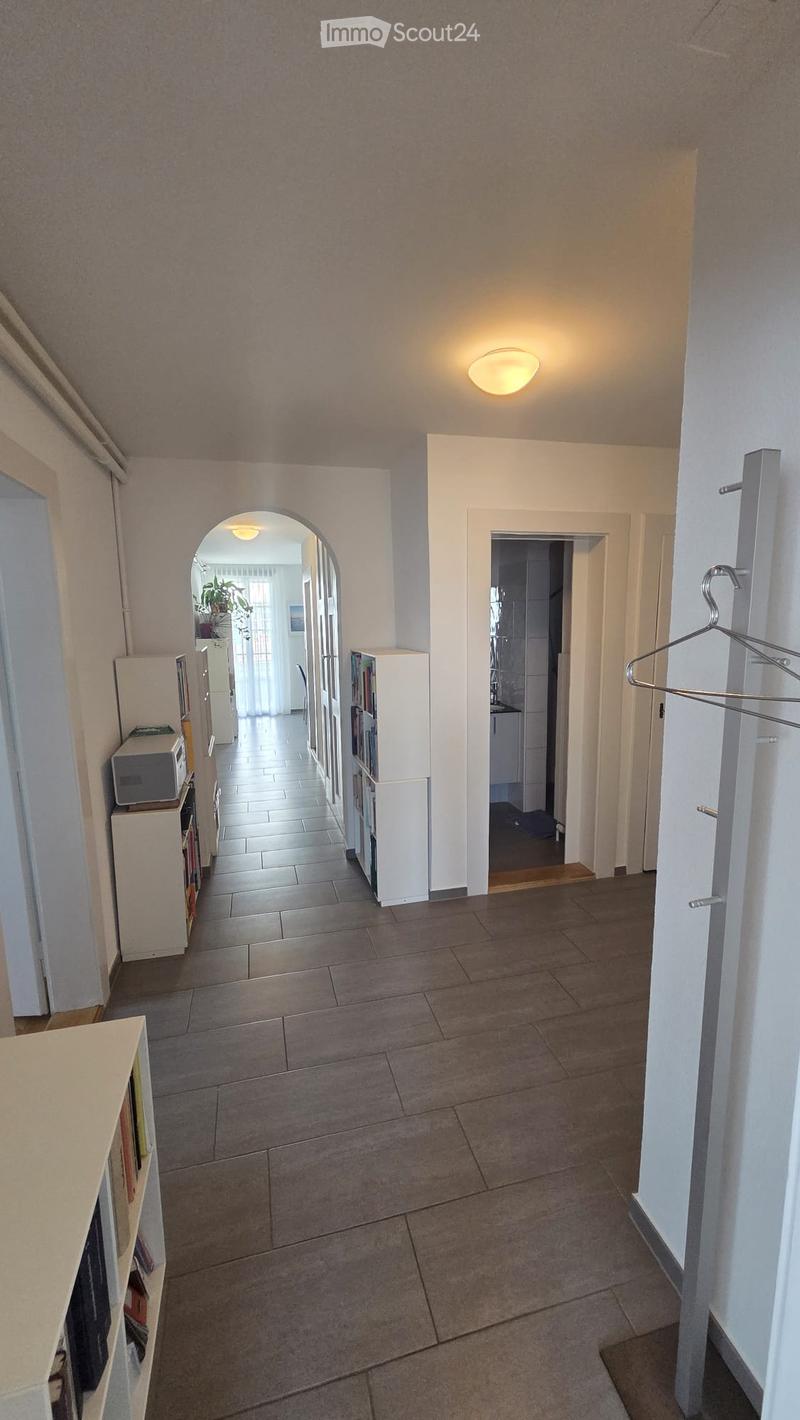 A well-lit hallway with tiled flooring, white walls, and several doors leading to different rooms. A coat rack is visible on the right side, and a small shelf with books and a microwave is on the left.