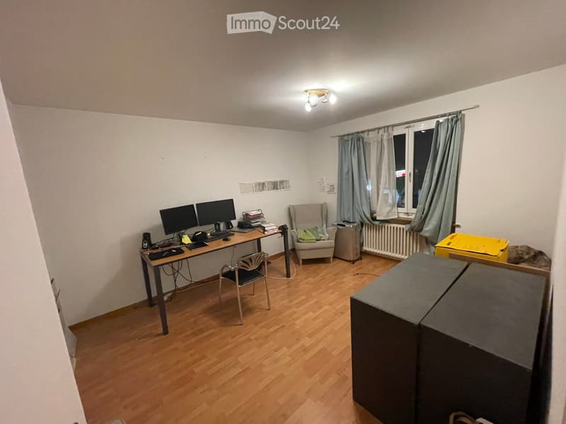 A furnished room with a wooden floor, a desk with two screens, a chair, a sofa, and a radiator near the window.