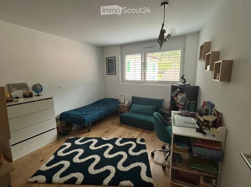 Cozy, well-lit room with a wooden floor, a blue sofa, a single bed, a desk with a chair, a bookshelf, and decorative items including a globe and a picture frame.