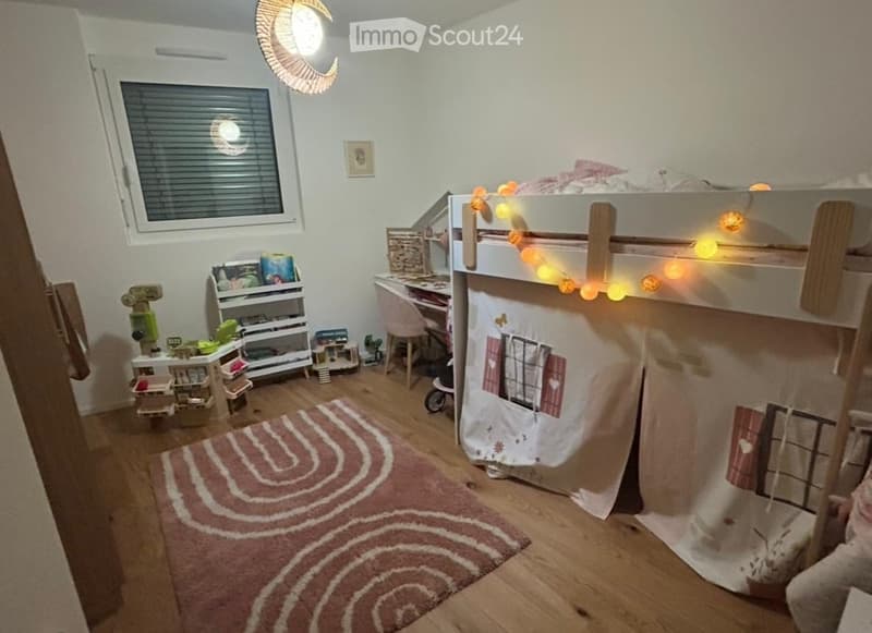 Cozy children's bedroom with a lofted bed, desk, shelves, and toys. Features a patterned rug and string lights.