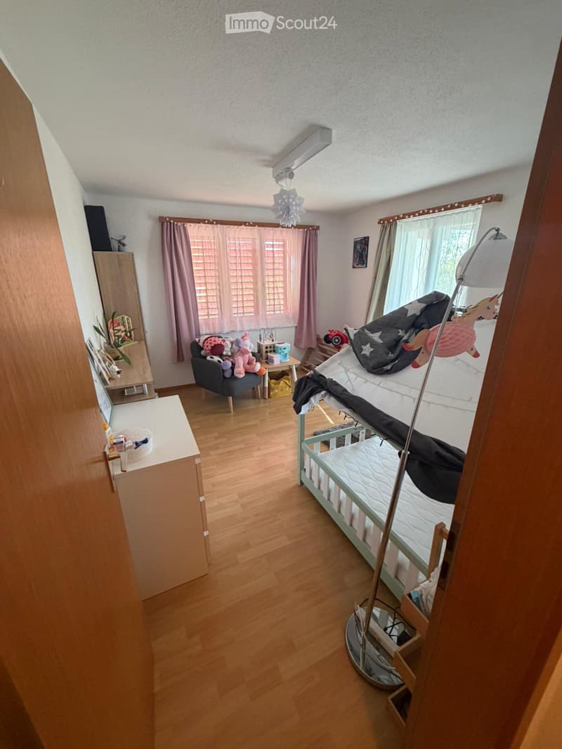 A well-lit bedroom with wooden flooring, a bunk bed, a small sofa with stuffed toys, a side table, a mirror, and a lamp.