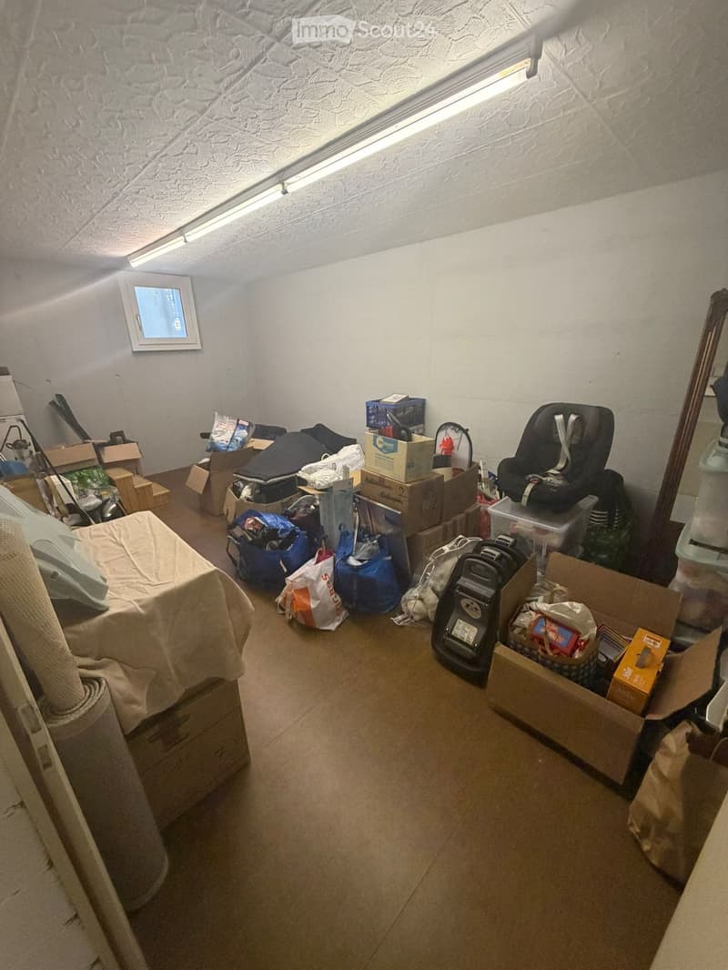 The room is cluttered with various items including a bed, a chair, storage boxes, a vacuum cleaner, a baby chair, and several bags and boxes scattered around.