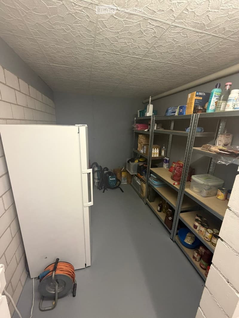 Storage room with a refrigerator, metal shelving units filled with various items, cleaning supplies, and a cable reel on the floor.
