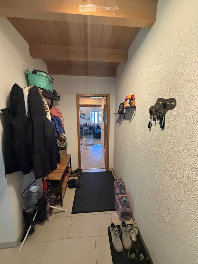 A narrow hallway with tiled flooring, wooden ceiling beams, and a black runner. Shoes and jackets are placed on shelves and racks along the walls, with a small table and a vase visible in the background.