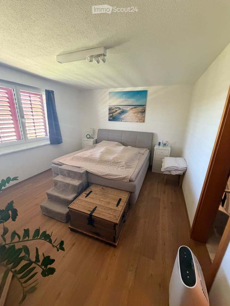 Cozy bedroom with a wooden floor, a large bed with a grey headboard, a wooden chest at the foot of the bed, a nightstand with a lamp, a painting on the wall, and a fan on the floor.