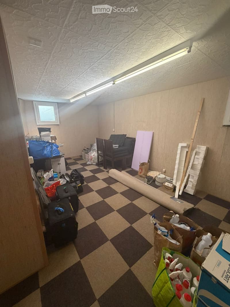 A cluttered room with a checkered floor, a chair, a table, and various items including suitcases, bags, and boxes.