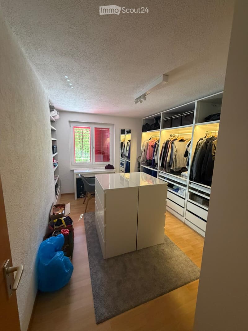 Well-lit room with wooden flooring, large built-in wardrobe with hanging clothes, a small table, and a chair near the window.