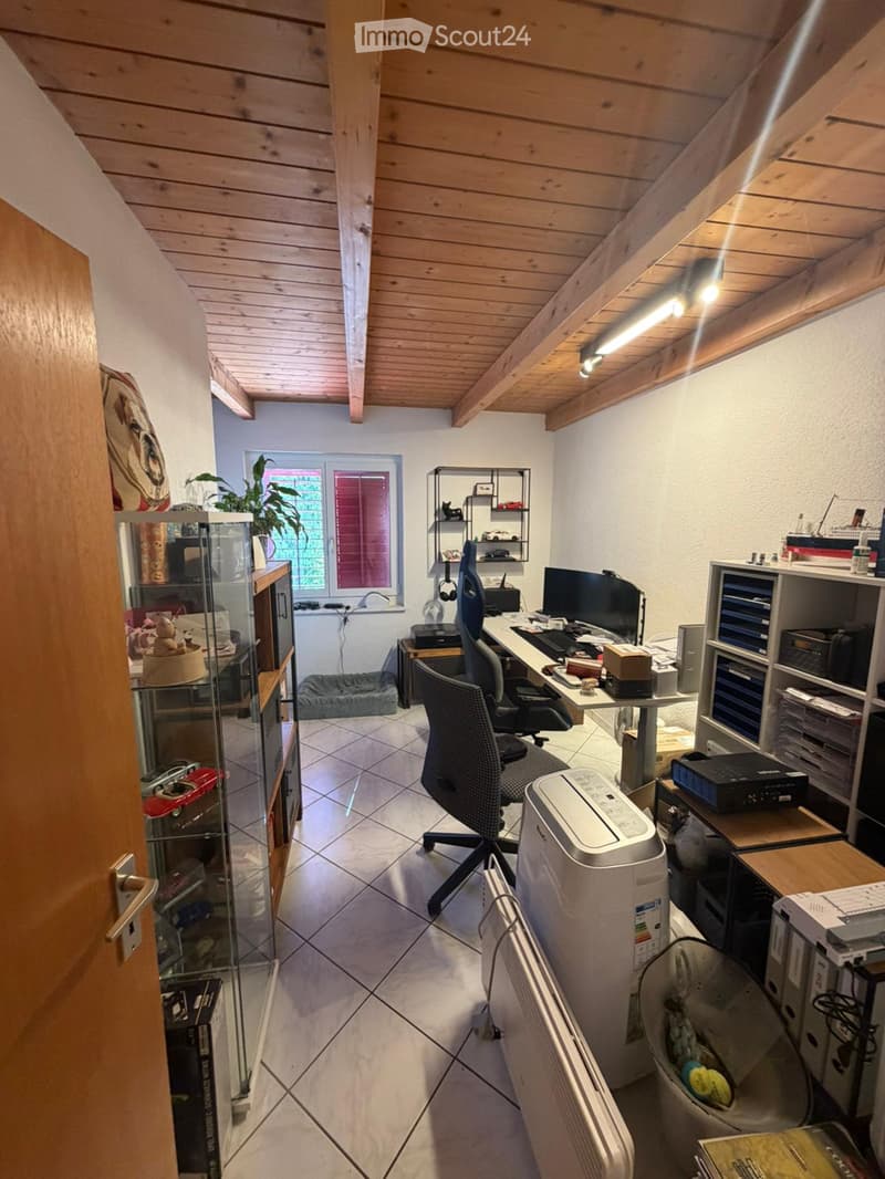 A well-lit, furnished room with a wooden slanted ceiling, tiled floor, and large window. Contains a desk with a computer, office chair, printer, shelves with books and decorative items, a cat bed, and a potted plant.