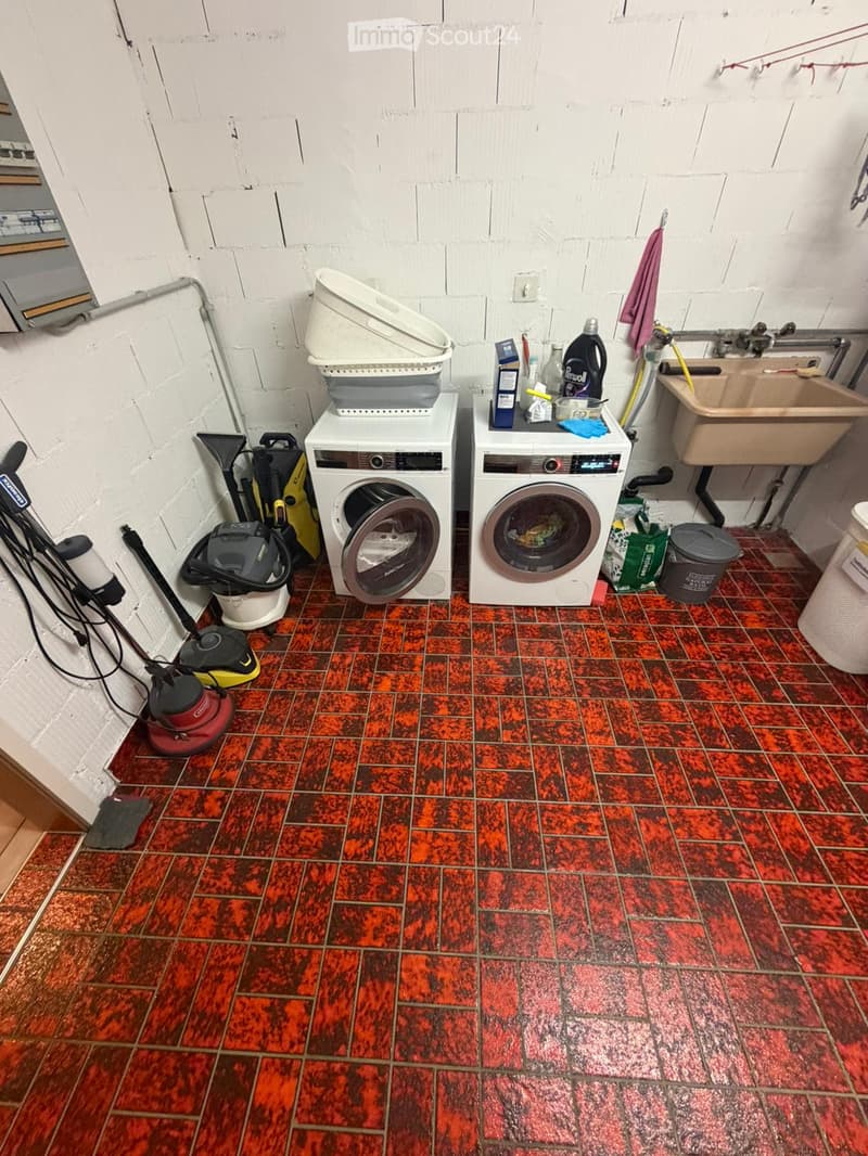 Bathroom with red and black tiled floor, white brick walls, washing machine, dishwasher, sink, cleaning supplies, and vacuum cleaner.