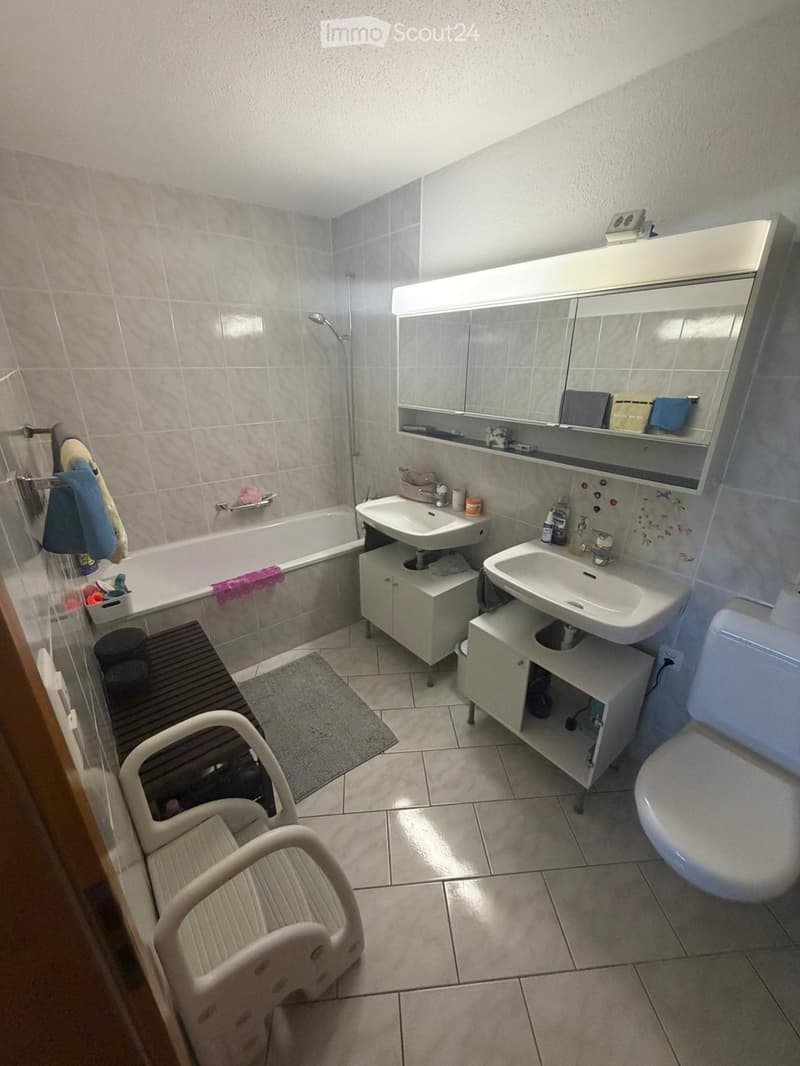 Bathroom with a bathtub, two sinks, a toilet, a chair, a mirror, and a cabinet. The room features tiled walls and floor, with towels and a small rug visible.