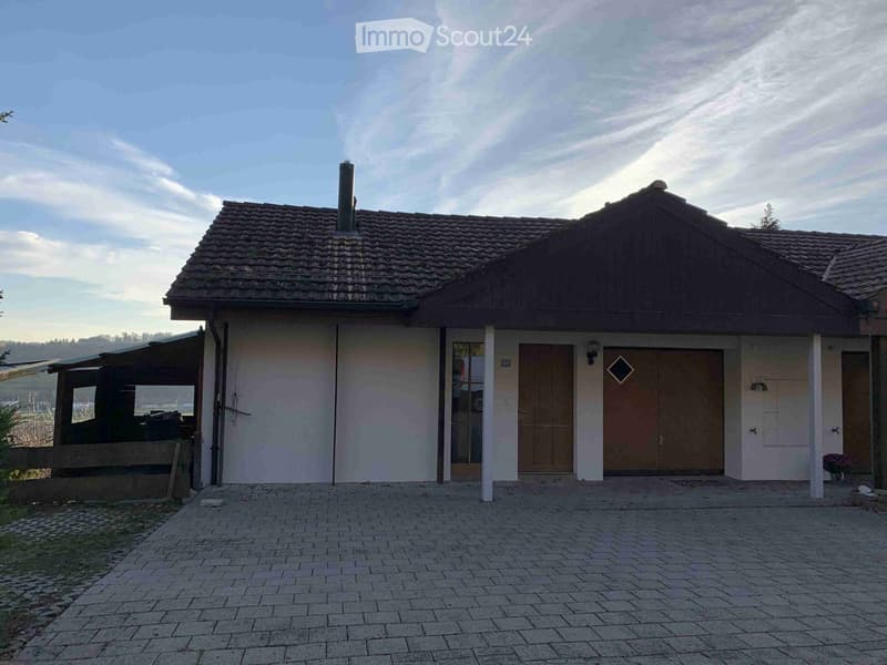 Two-story house with a tiled roof, white walls, and a covered porch area. Features include a garage door, a main entrance door, and a small garden area with a potted plant.