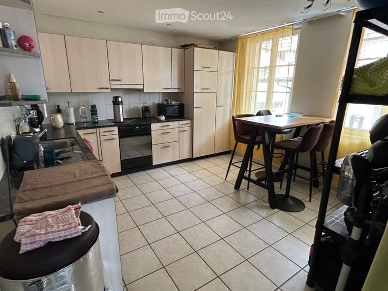 A modern kitchen with light-colored cabinets, a black countertop, and a small dining area with a table and chairs. The kitchen features various appliances and a tiled floor.