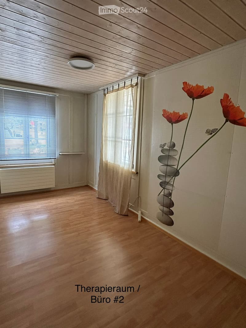Empty room with wooden flooring, large window with blinds, radiator, and a decorative flower arrangement with stones on a stand.