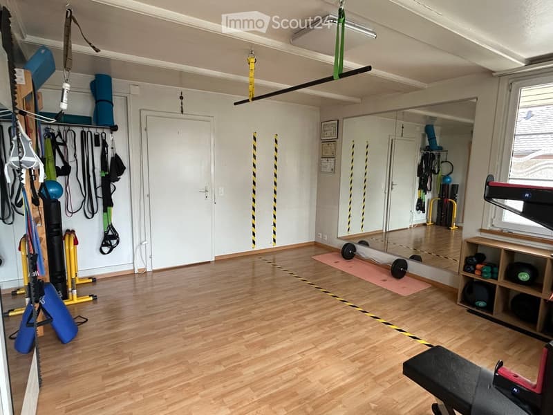 Well-lit room with wooden flooring, large mirror, fitness equipment including weights, balls, and a shelf with additional gear.
