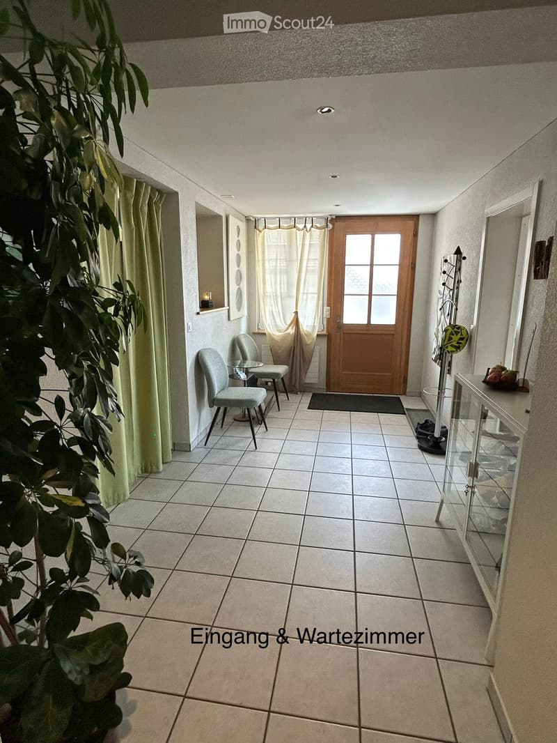 Entrance and waiting area with tiled floor, two chairs, small table, coat rack, and a large plant on the left side.