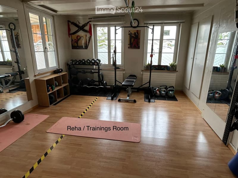 Well-lit room with wooden flooring, multiple windows, and various fitness equipment including weights, a weight bench, kettlebells, shelves, and yoga mats.
