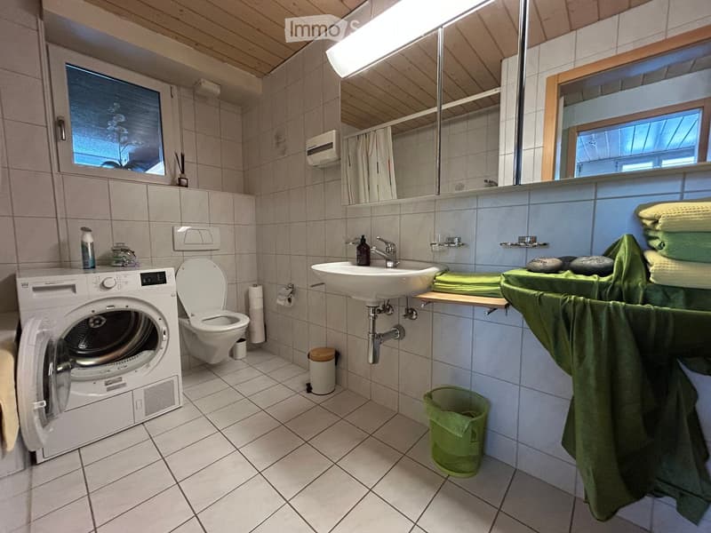 Bathroom with a washing machine, toilet, sink, mirror, and neatly stacked towels. The room features tiled walls and floor, with a window providing natural light.